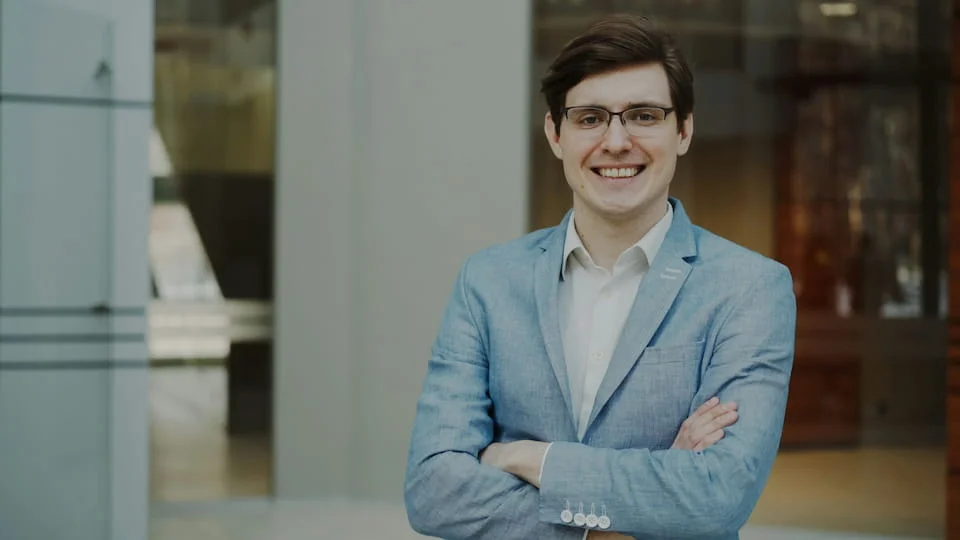 A man in a light blue suit wearing glasses, smiling.